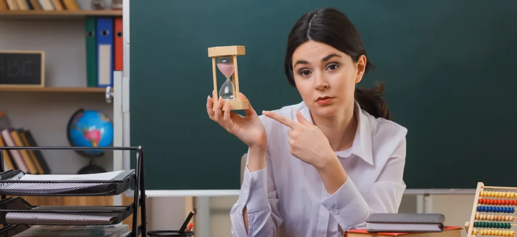 A teacher sitting at a desk in a classroom holding an hourglass and pointing at it to emphasize the importance of time