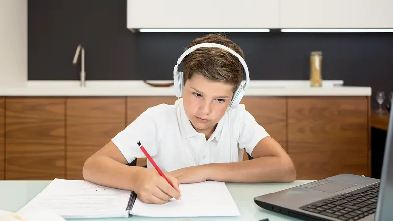 A young boy wearing headphones writing in a notebook while studying in front of a laptop at home
